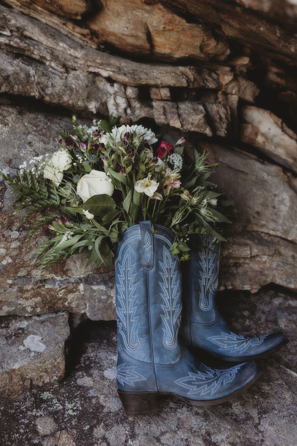 Blue cowboy boots filled with wildflower bouquet resting against rustic stone wall wedding detail.