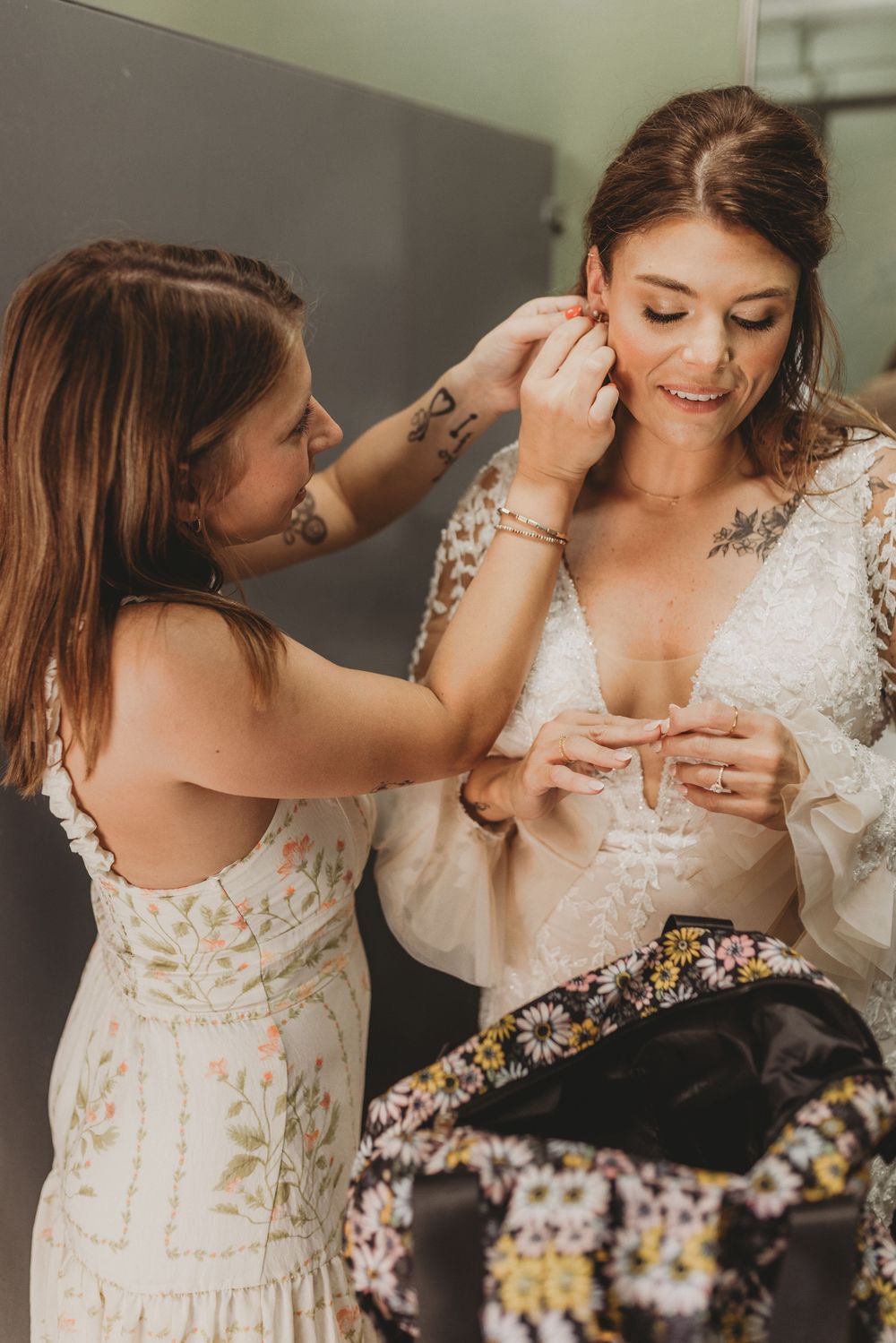 Bridesmaid helps smiling bride put on earrings while getting ready in bridal lace gown.