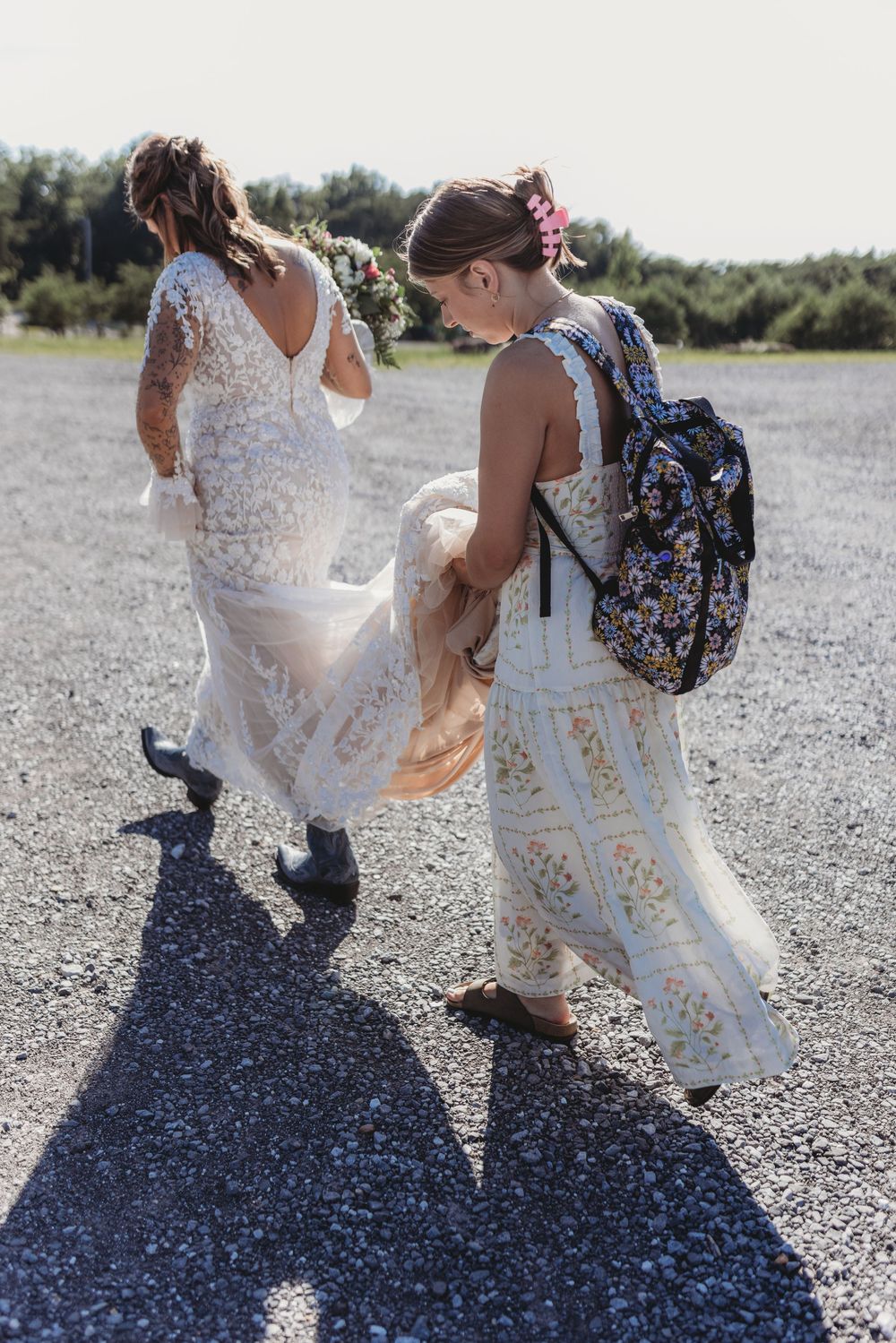 Bride in boho lace dress with flower crown walks with bridesmaid on gravel path in bright sunlight.