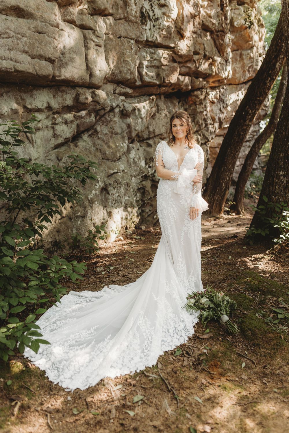 Bride in elegant lace wedding gown with long train poses near rustic stone wall surrounded by lush greenery.