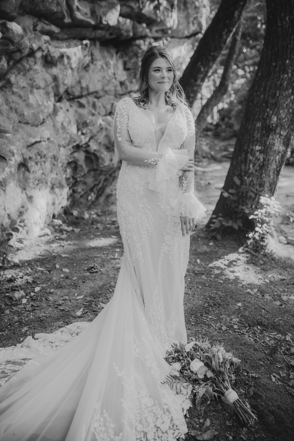 Black and white portrait of bride in lace mermaid gown with bell sleeves standing near rocky forest backdrop.