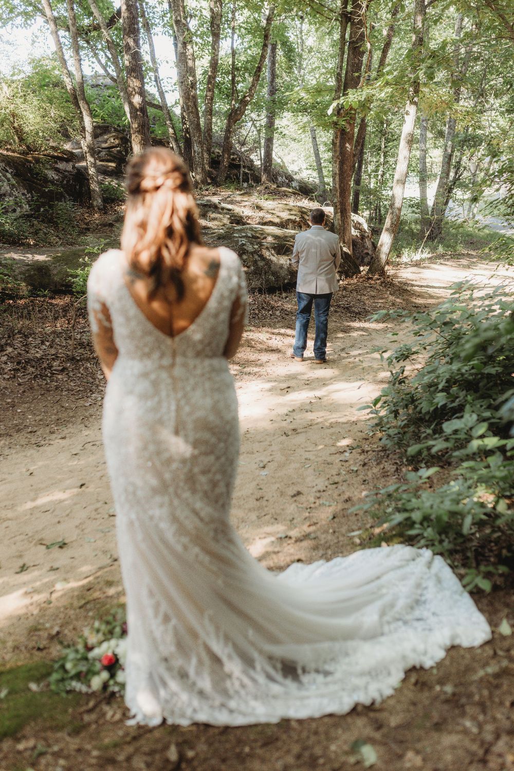 Bride in lace gown with long train approaching groom standing on wooded forest path for first look.