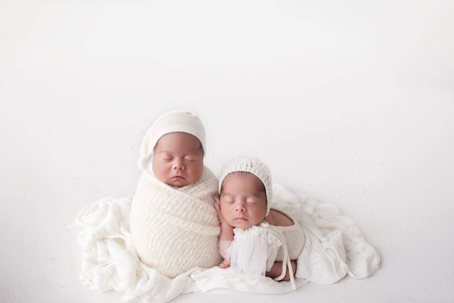 Two sleeping newborn twins wrapped in white swaddle blankets held by parents hands on white background.