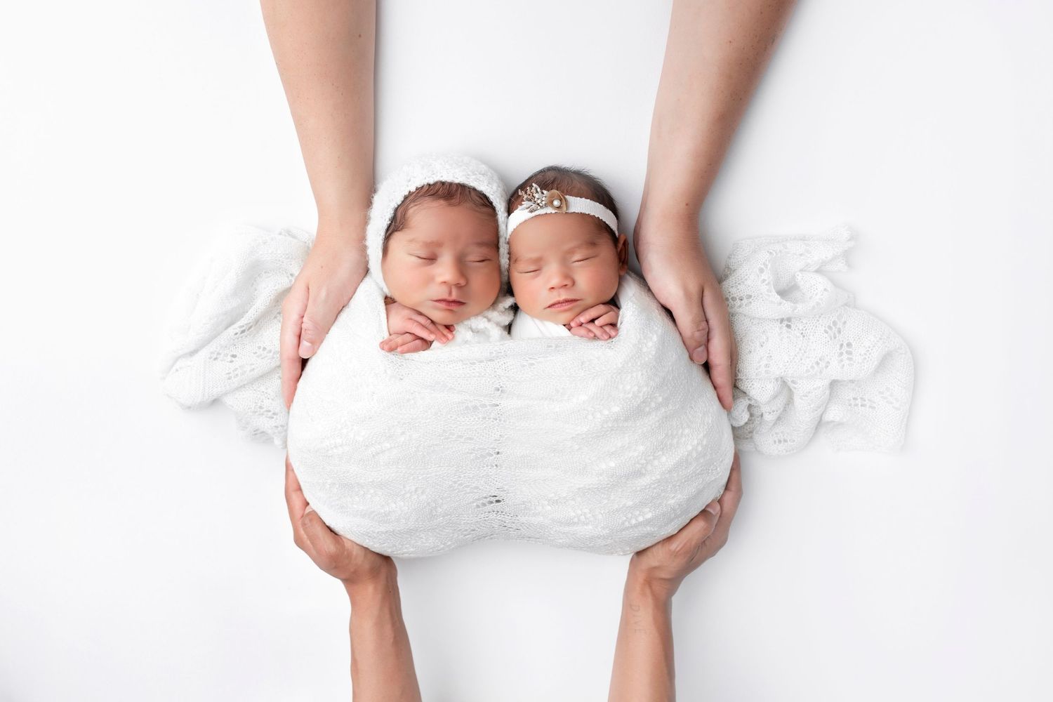 Two sleeping newborn twins wrapped in white swaddle blankets held by parents hands on white background.