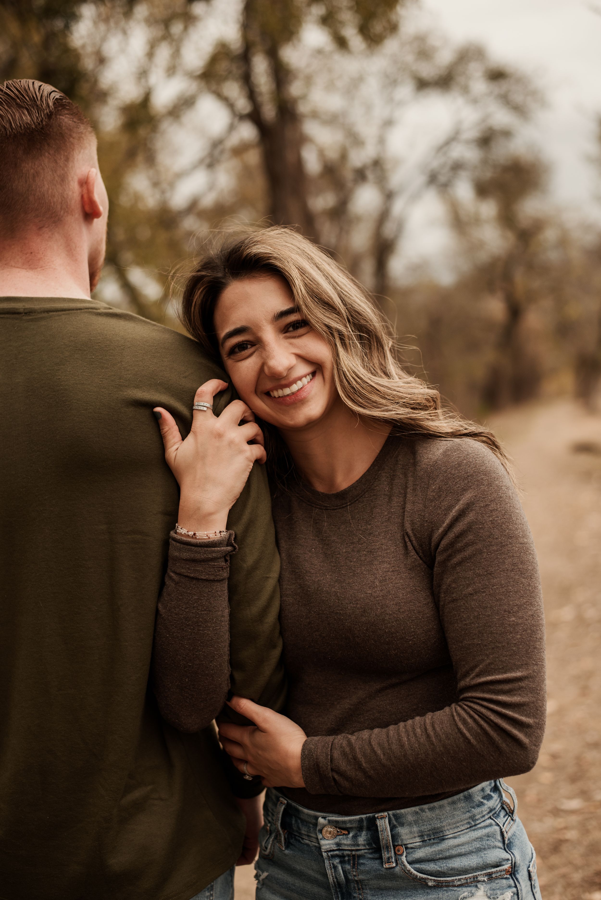 Smiling woman in brown sweater leaning against mans shoulder during outdoor fall engagement photo session.