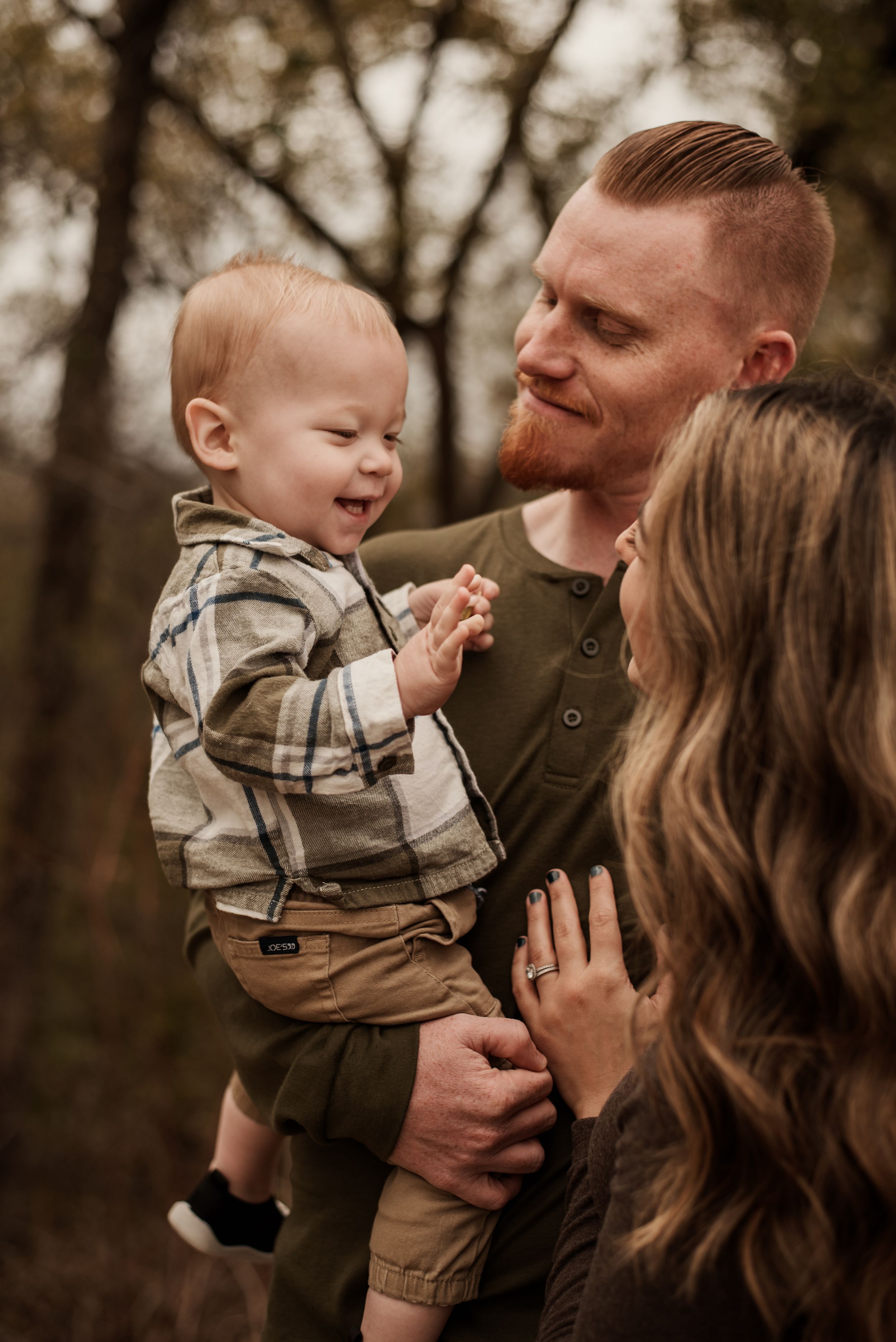 A red-haired father holds his laughing baby boy while a woman with wavy brown hair looks on during an outdoor family portrait.