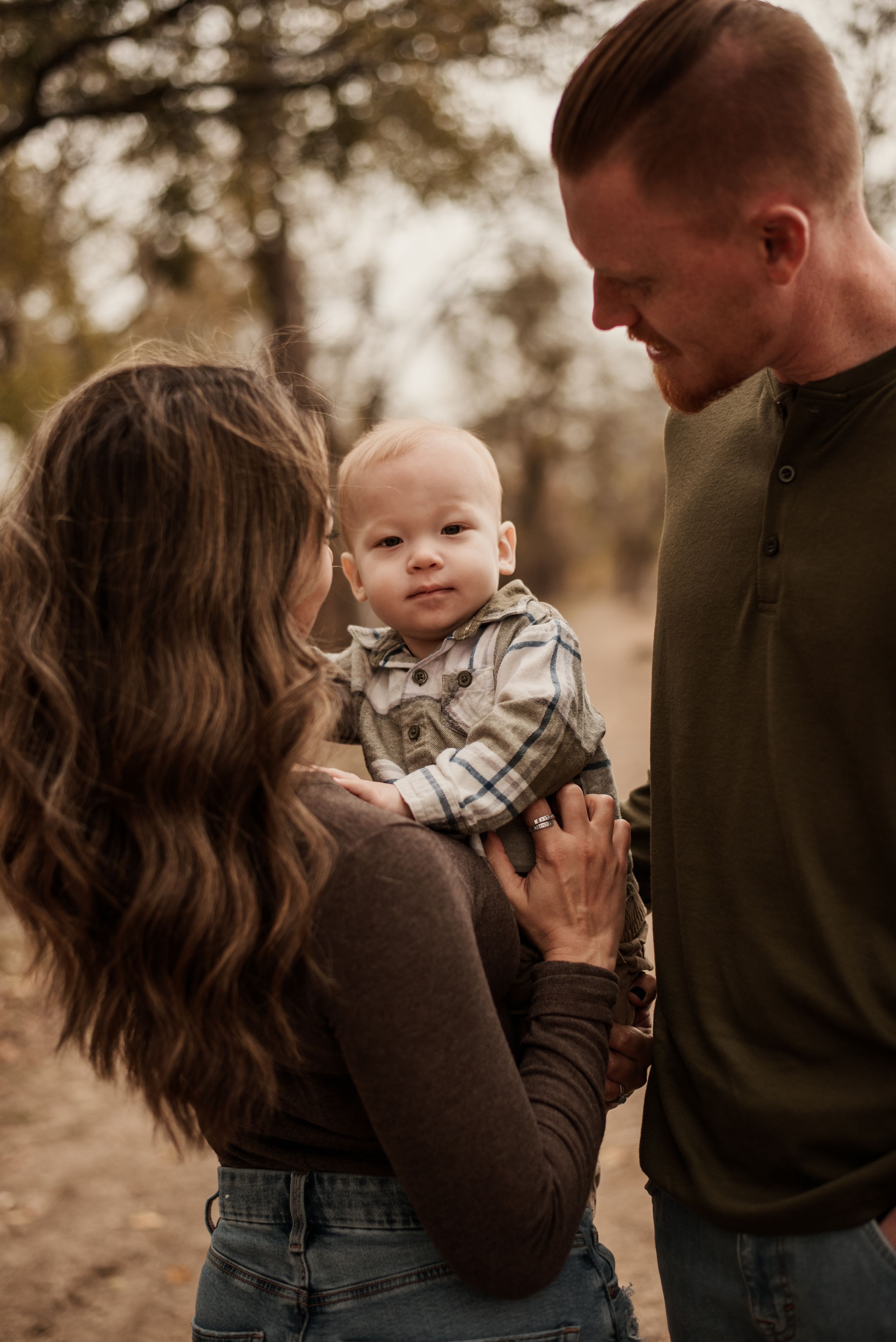 A young family with a baby boy enjoying an outdoor autumn portrait session in a wooded setting.