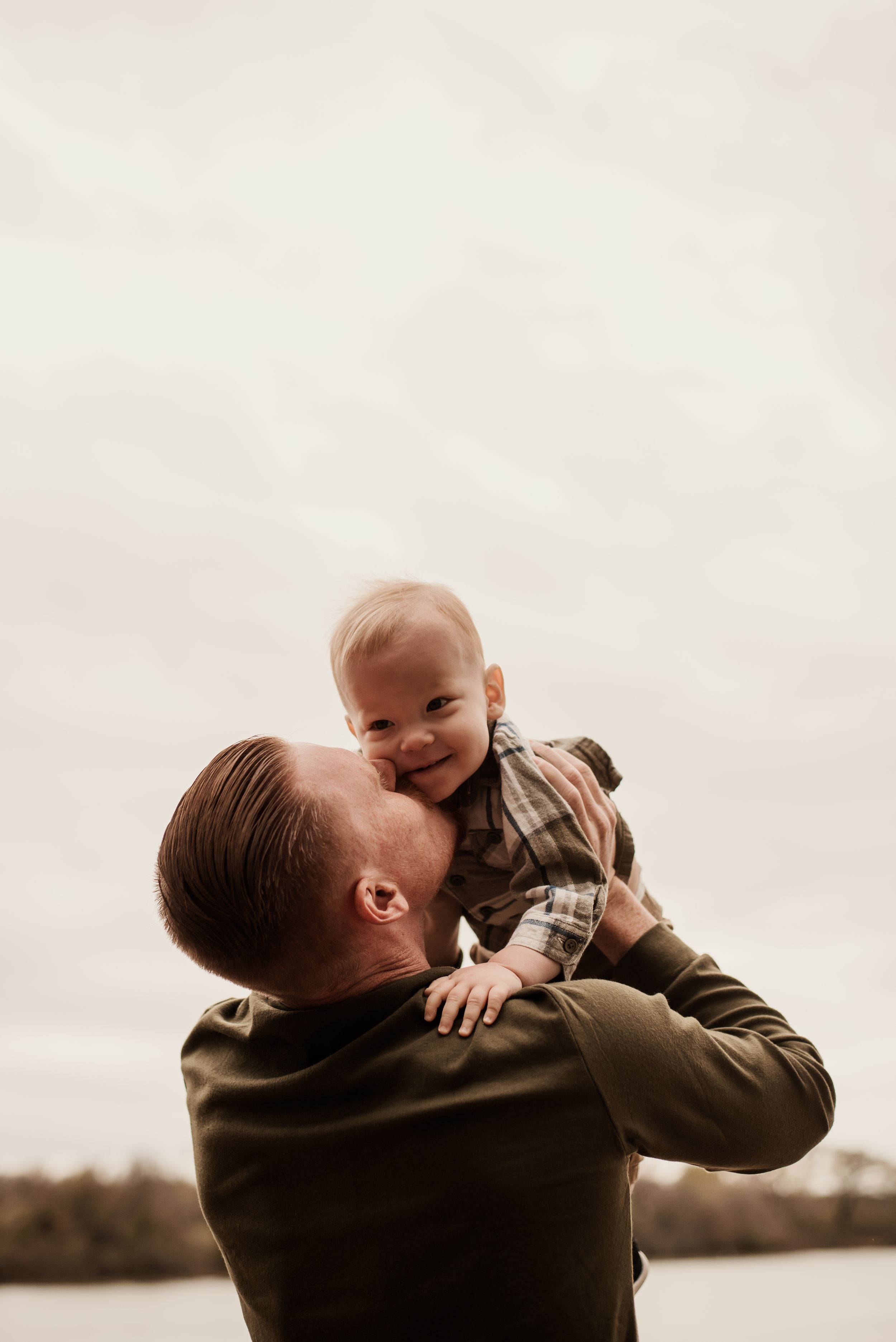 A father lovingly lifts his smiling baby boy overhead outdoors on a cloudy day.