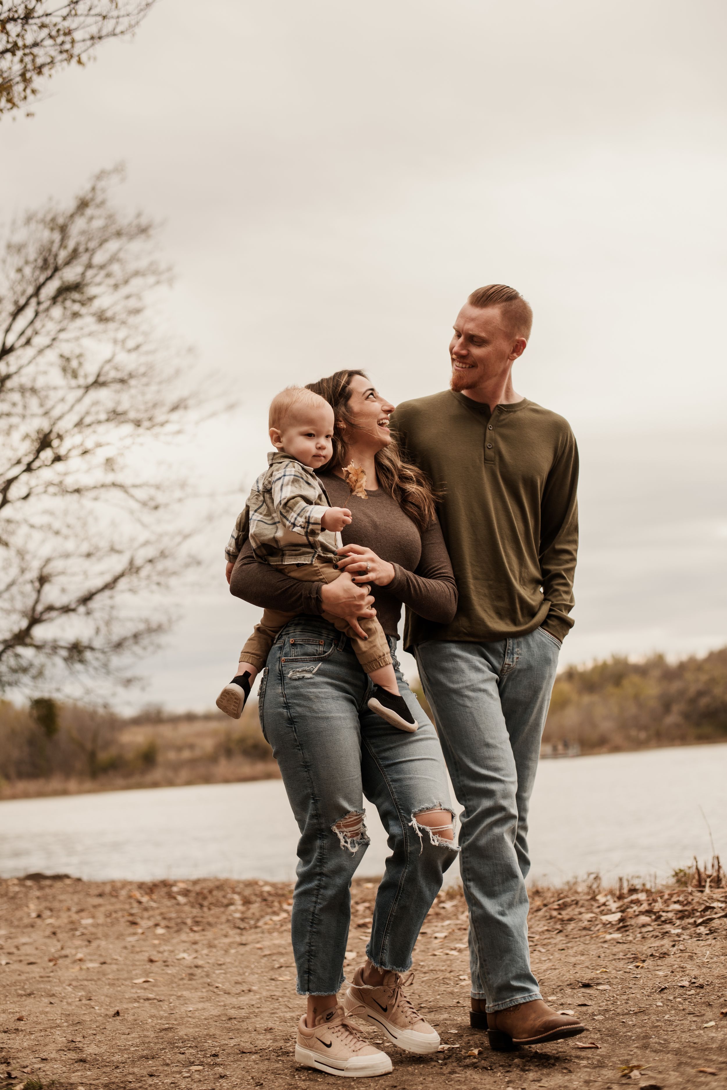 A young family with a baby poses outdoors near a lake in autumn, wearing casual jeans and earth-toned tops.