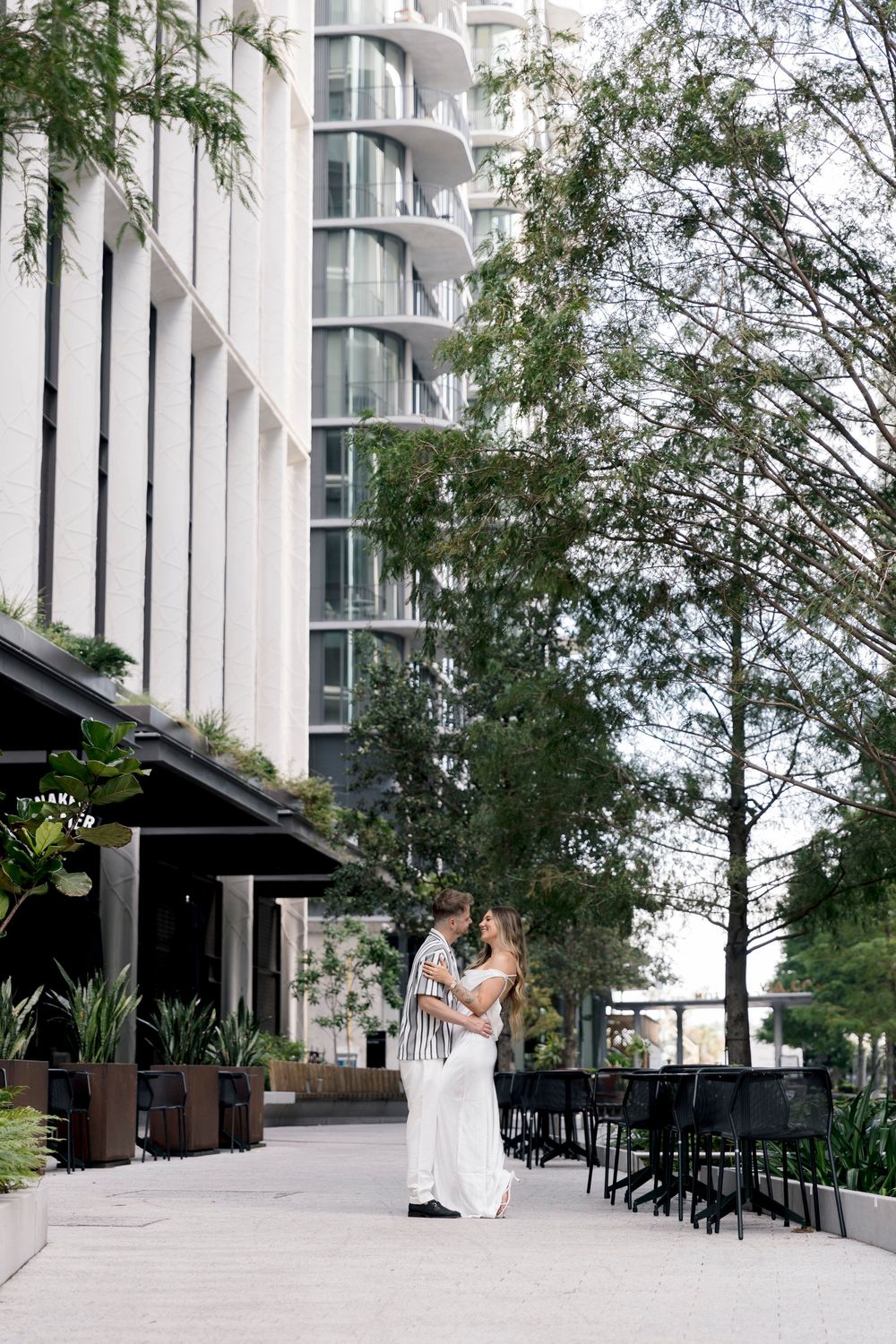 Couple embracing romantically outside a modern high-rise building surrounded by lush green trees and outdoor seating.