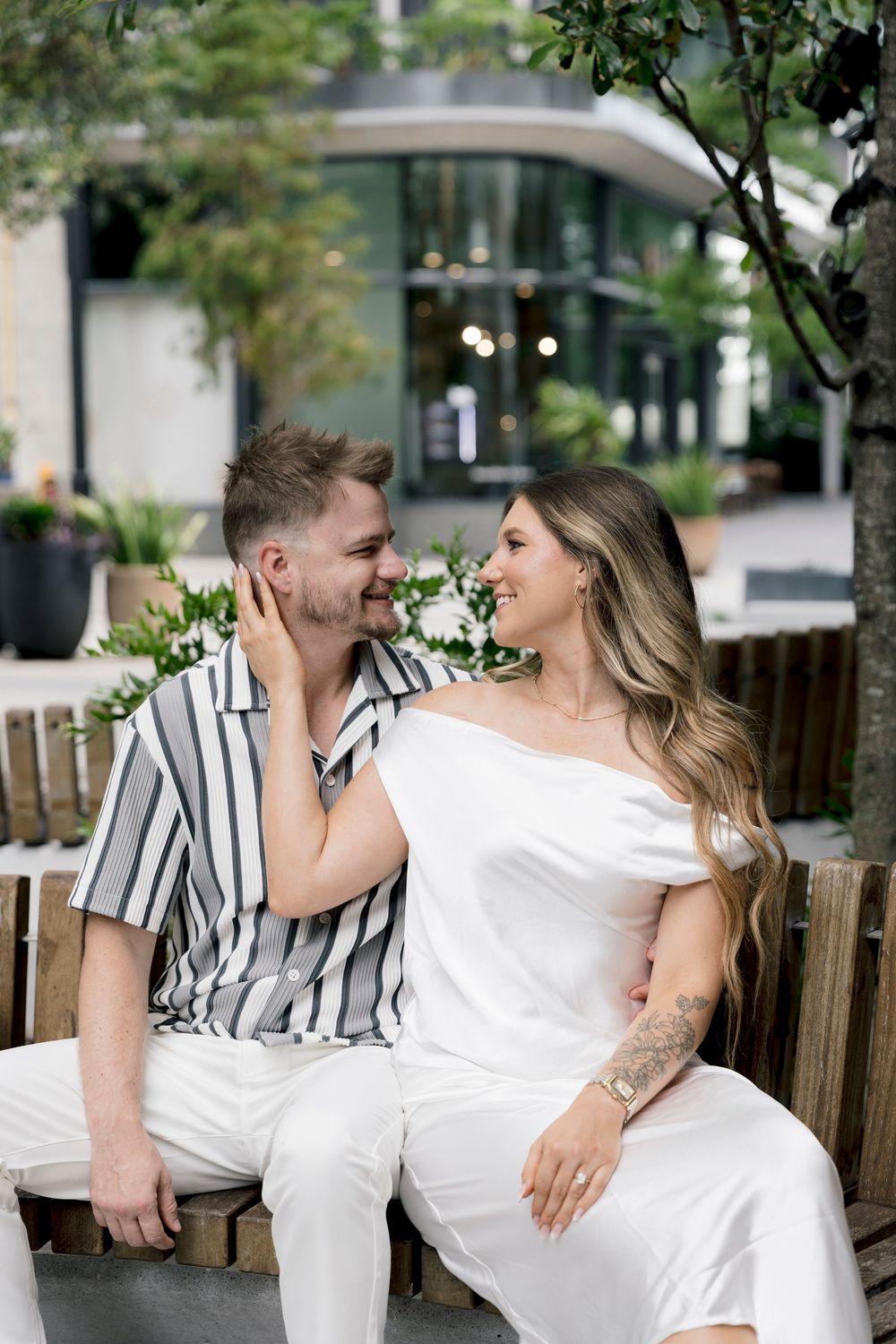 A smiling couple sits on a wooden bench outdoors, woman in white jumpsuit touching mans face, both gazing at each other.