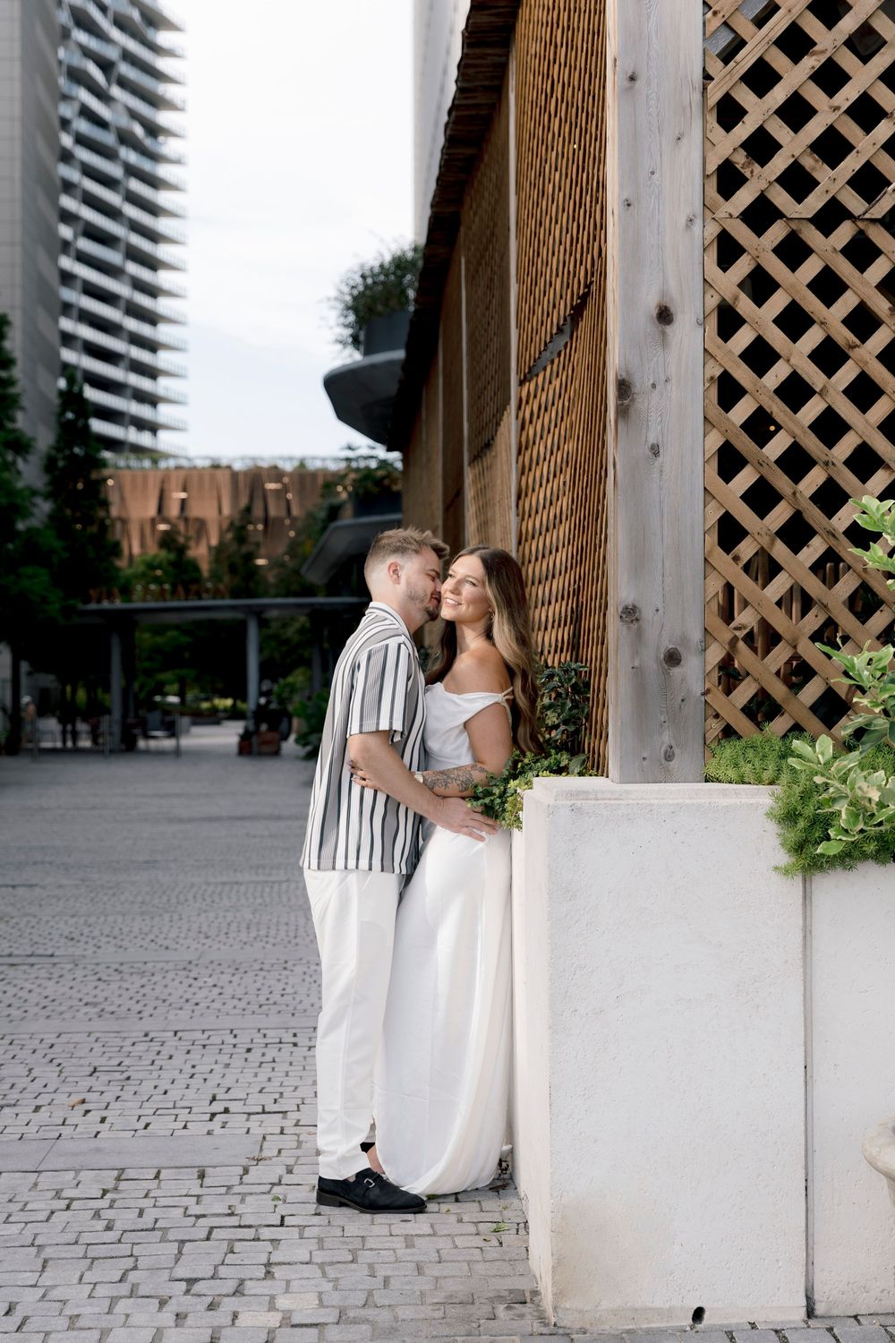 A couple shares a romantic embrace near a wooden lattice wall and planter in an urban plaza with modern buildings.
