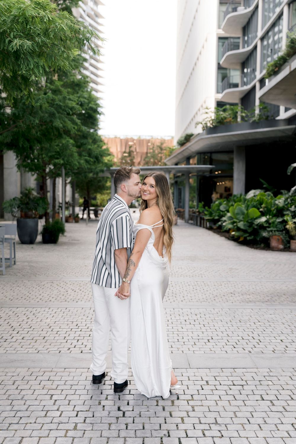 Couple in white outfits holding hands on a cobblestone plaza surrounded by modern buildings and lush greenery.