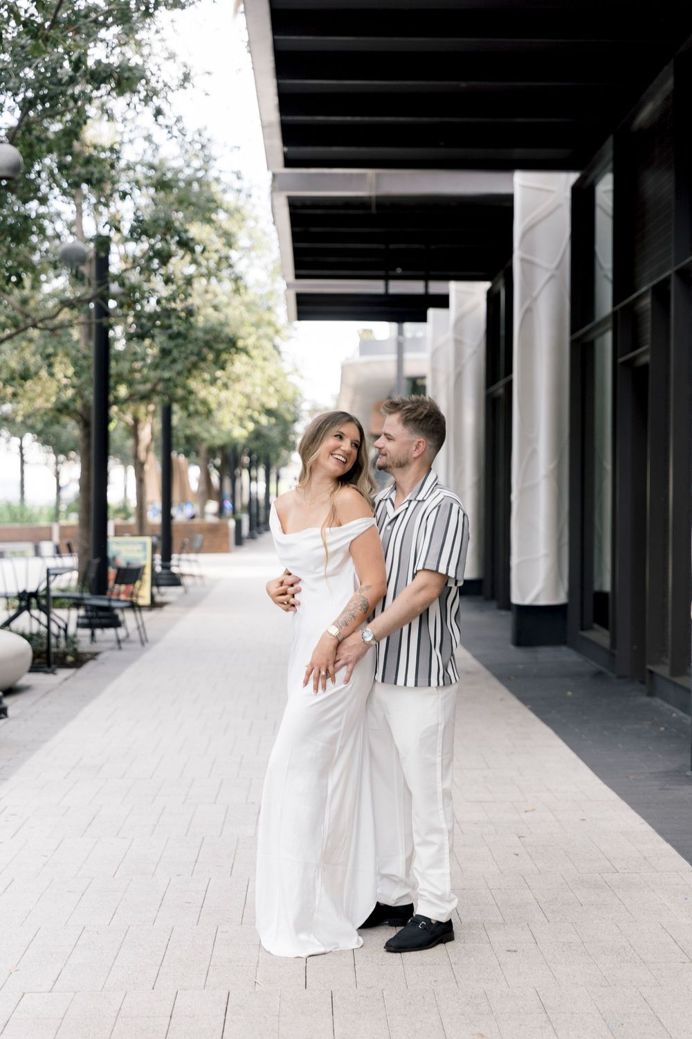 A couple embraces on an urban sidewalk, the woman in a white jumpsuit and the man in a striped shirt, sharing a joyful moment.