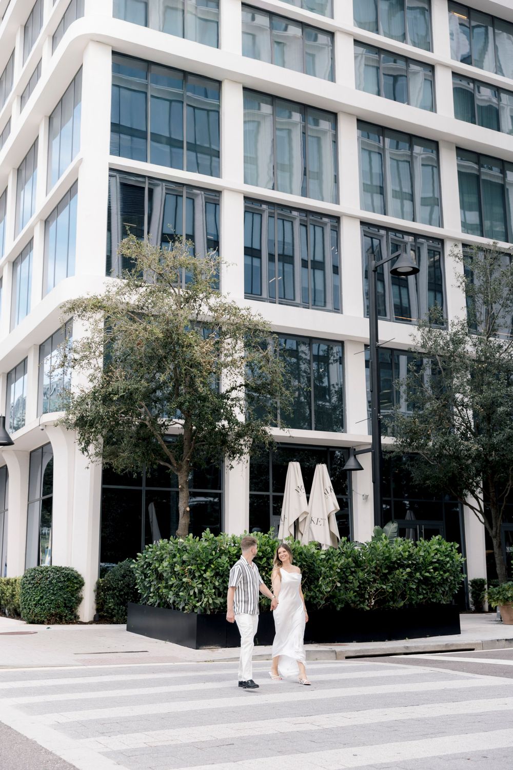 Couple holding hands crossing a zebra crossing in front of a modern glass office building with green hedges.
