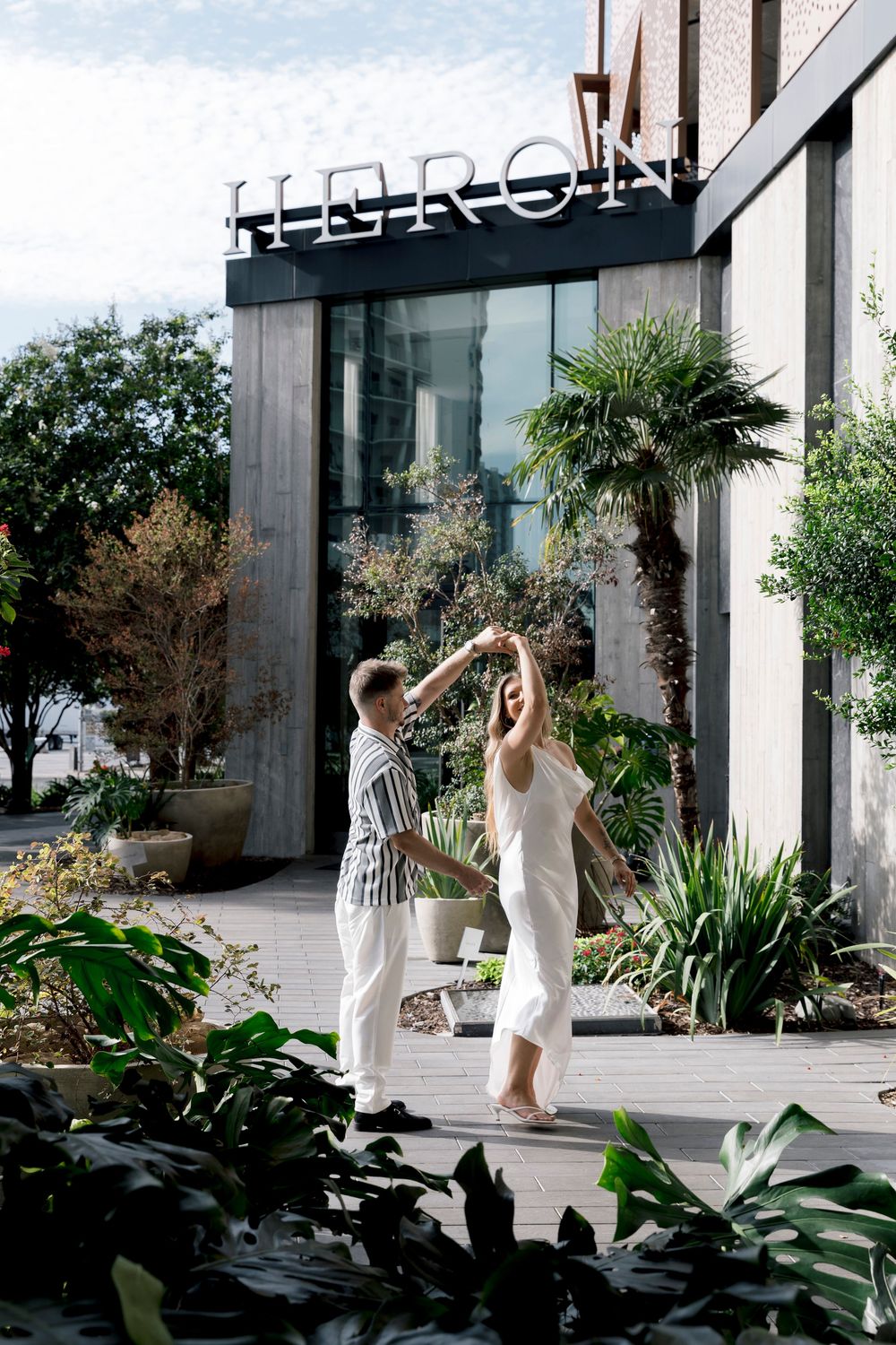 Couple dancing outdoors in front of Heron building, surrounded by lush tropical plants on a sunny day.
