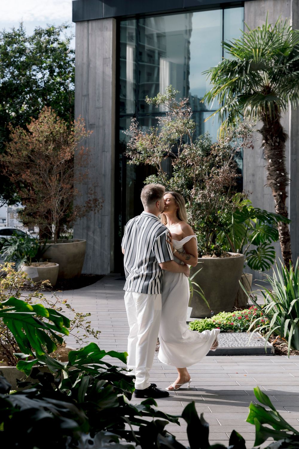 Romantic couple embracing and kissing on a sunlit patio with lush tropical greenery and glass building.