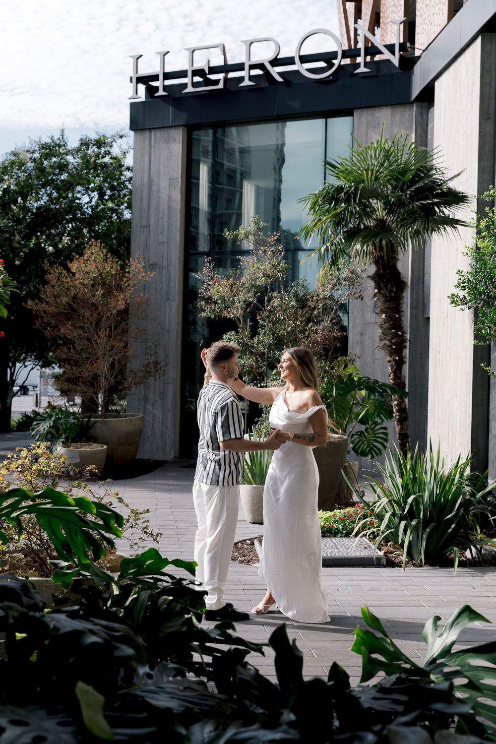 Couple dancing outside Heron restaurant surrounded by lush tropical plants and palm trees on a sunny day.