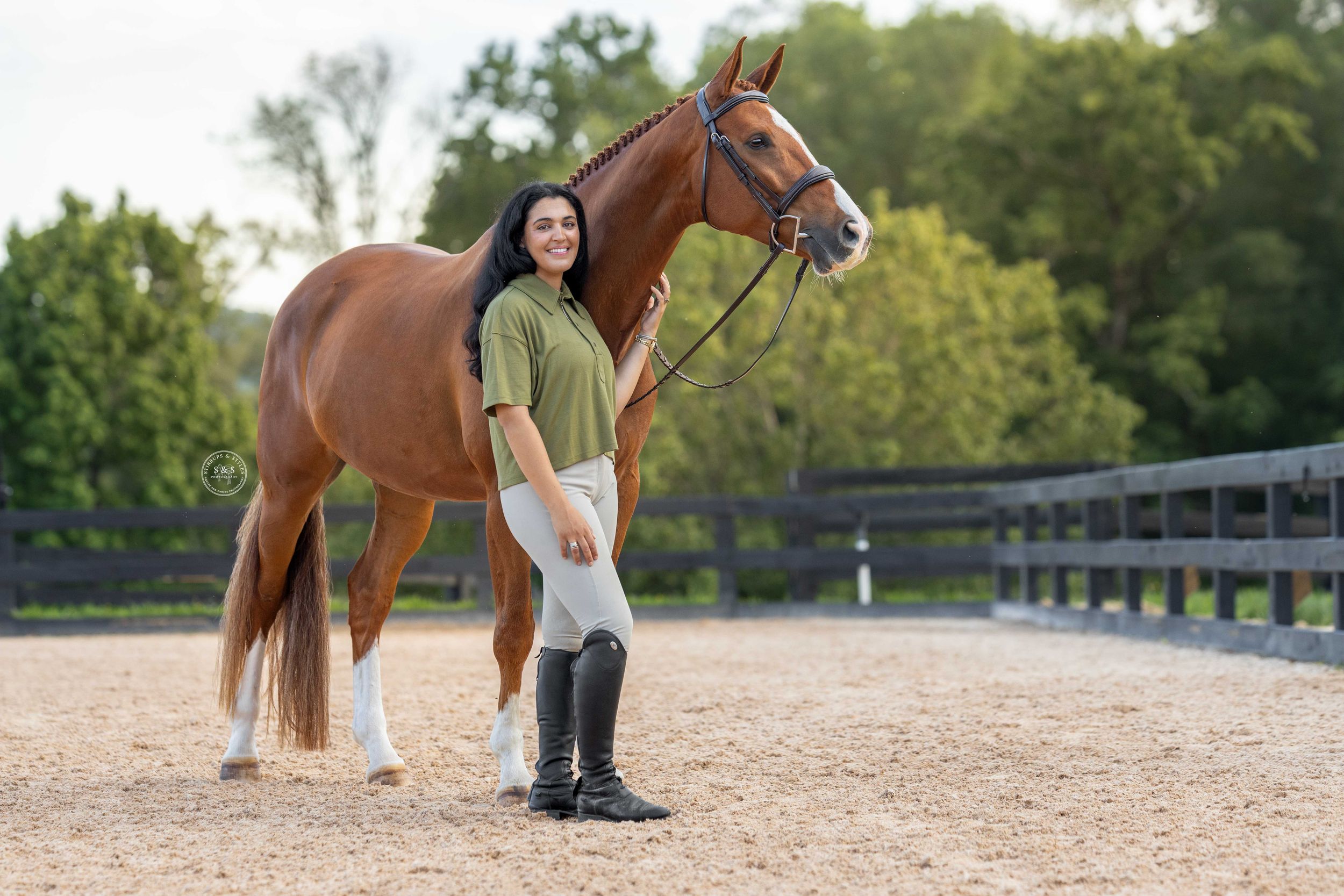 Woman in riding boots stands beside a chestnut horse in a sandy equestrian arena with green trees.