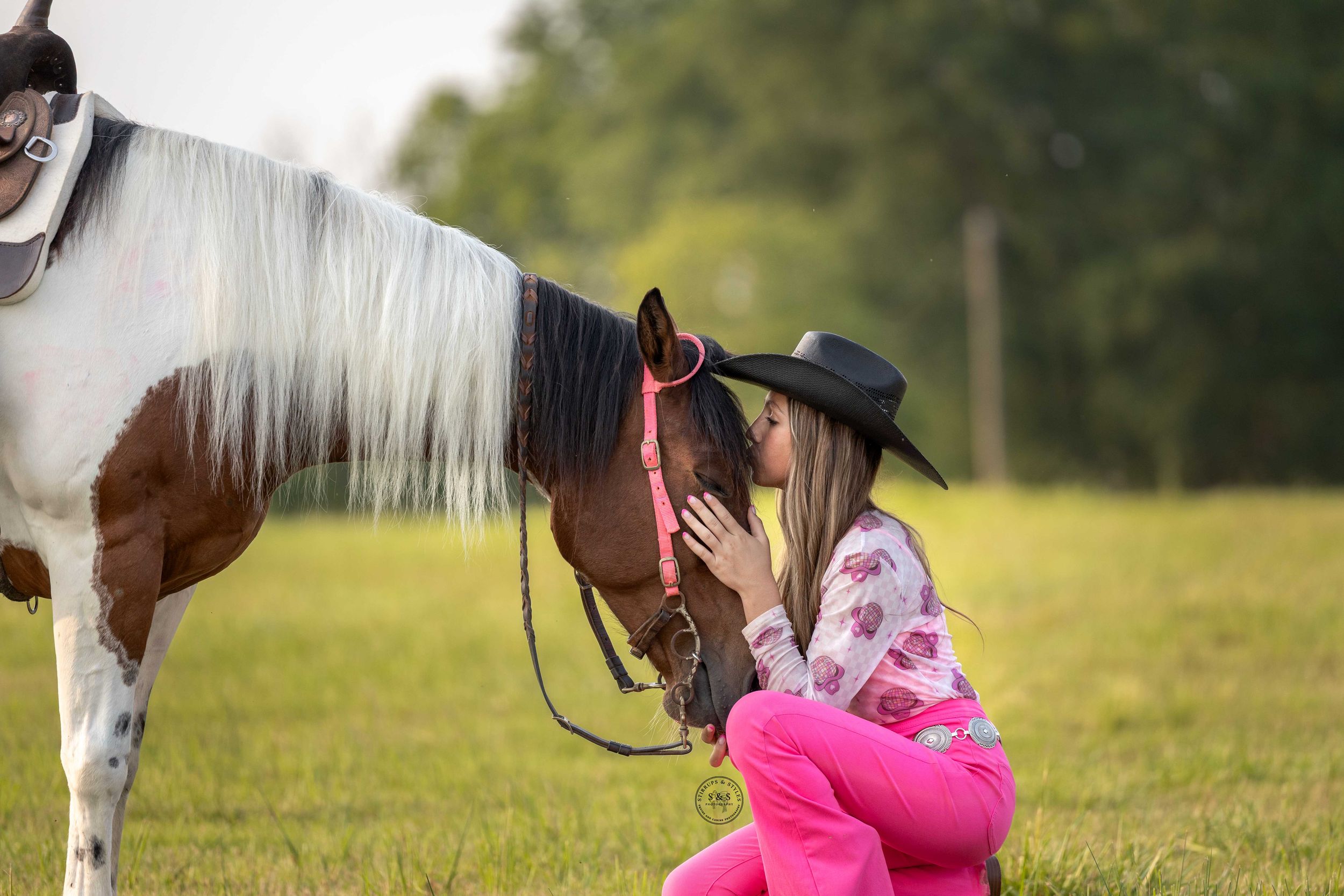 Young girl in pink outfit and black cowboy hat kneeling to kiss a paint horse on a green field.