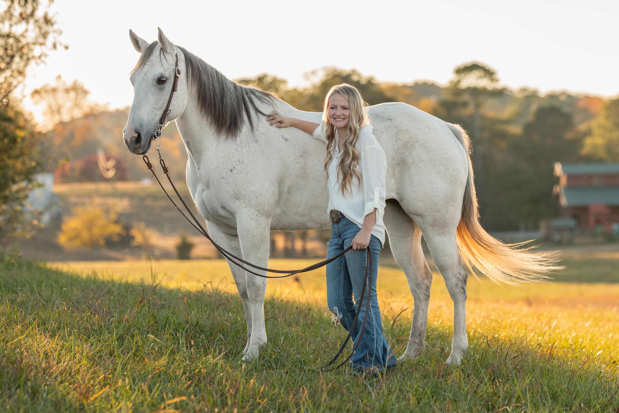Young blonde woman stands beside a white horse in a sunlit green pasture during golden hour.