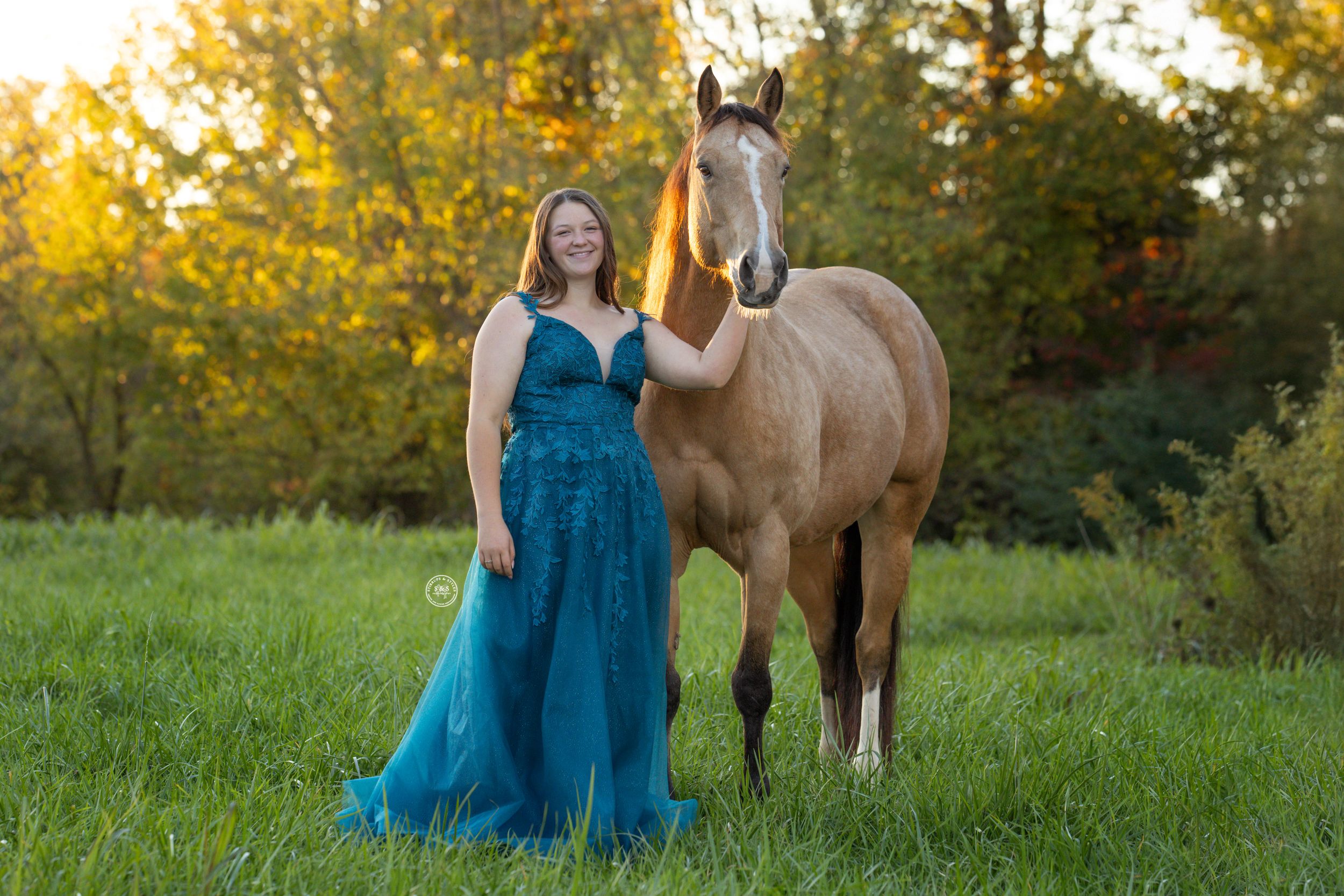 Woman in teal gown poses with buckskin horse in sunlit green meadow with autumn trees backdrop.