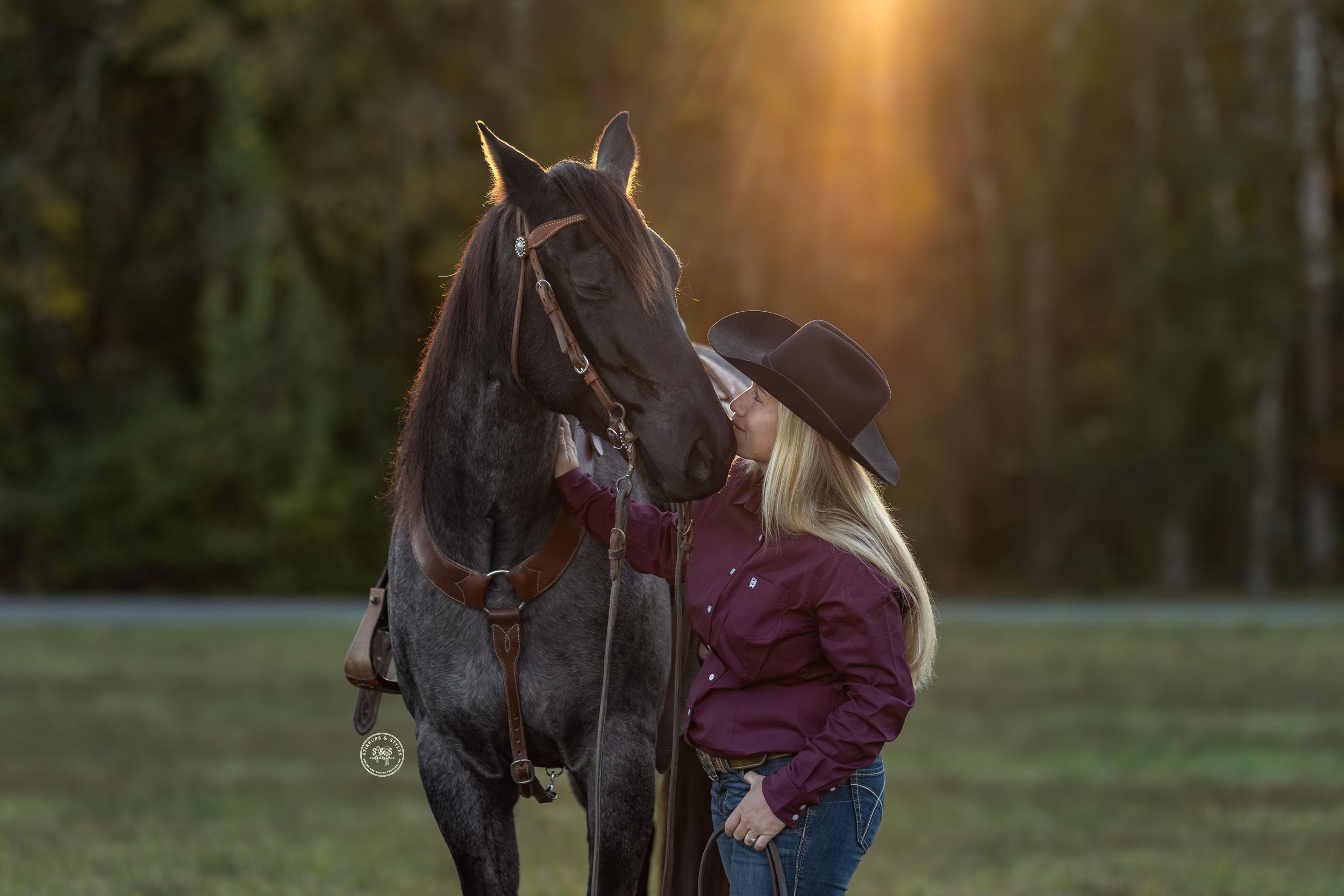 Blonde woman in cowboy hat sharing a tender moment with a dark horse at golden sunset in a field.