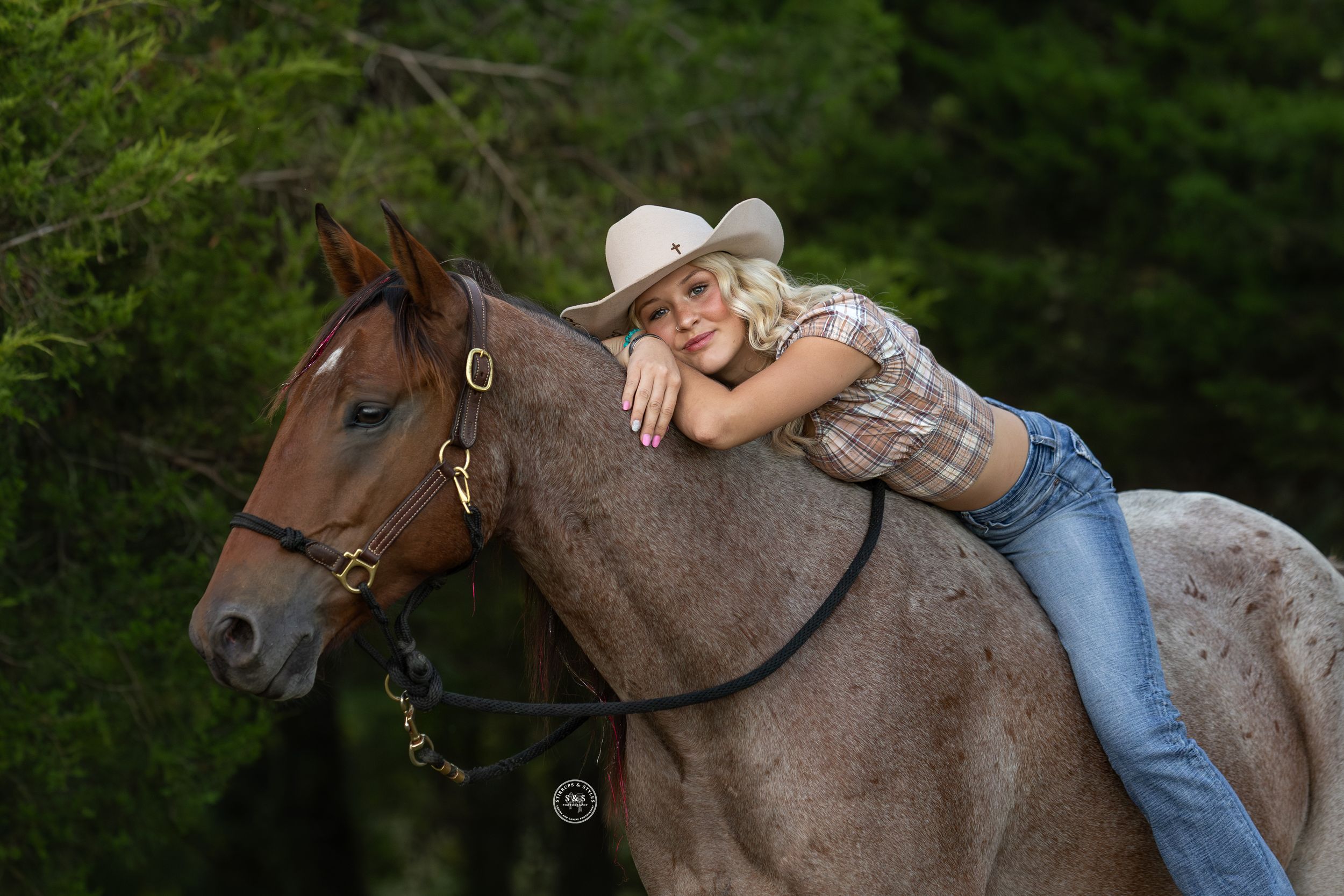 Blonde woman in cowboy hat and jeans leaning on a brown horse outdoors near green trees.