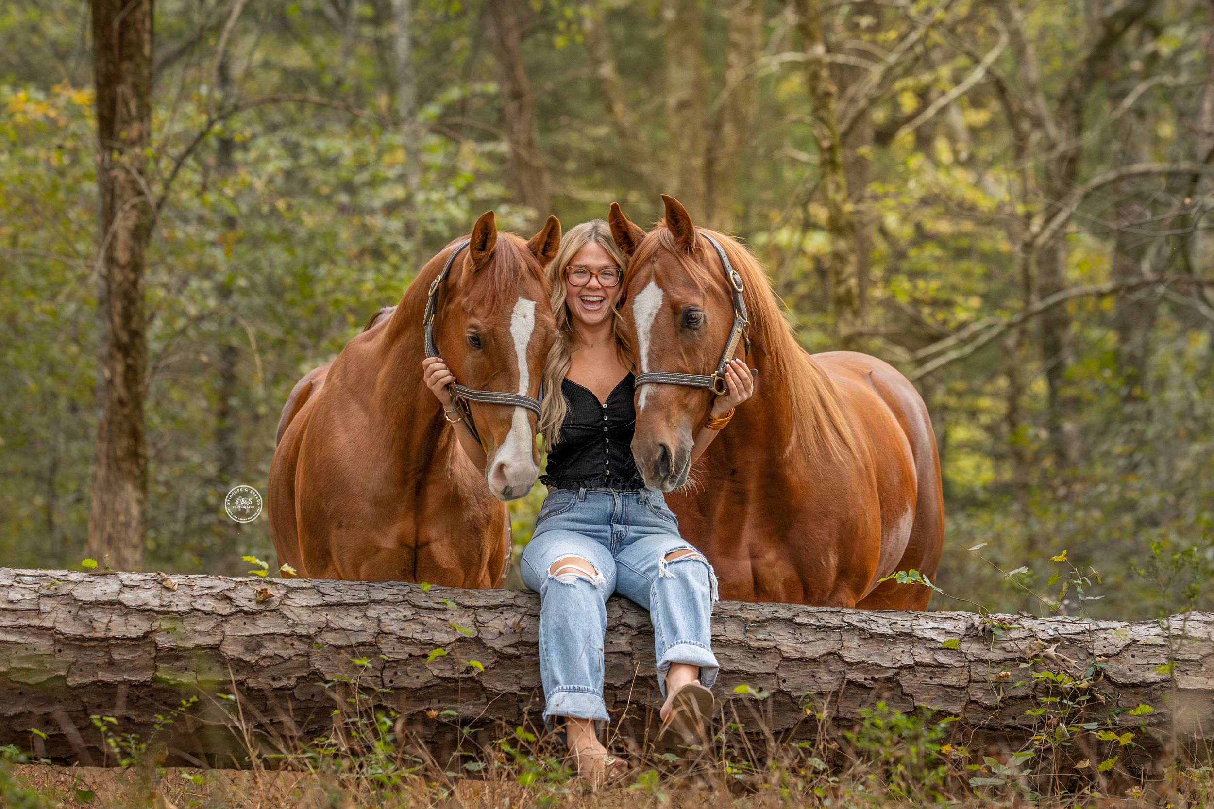 Young blonde woman sits on a log between two chestnut horses in a lush forest setting.