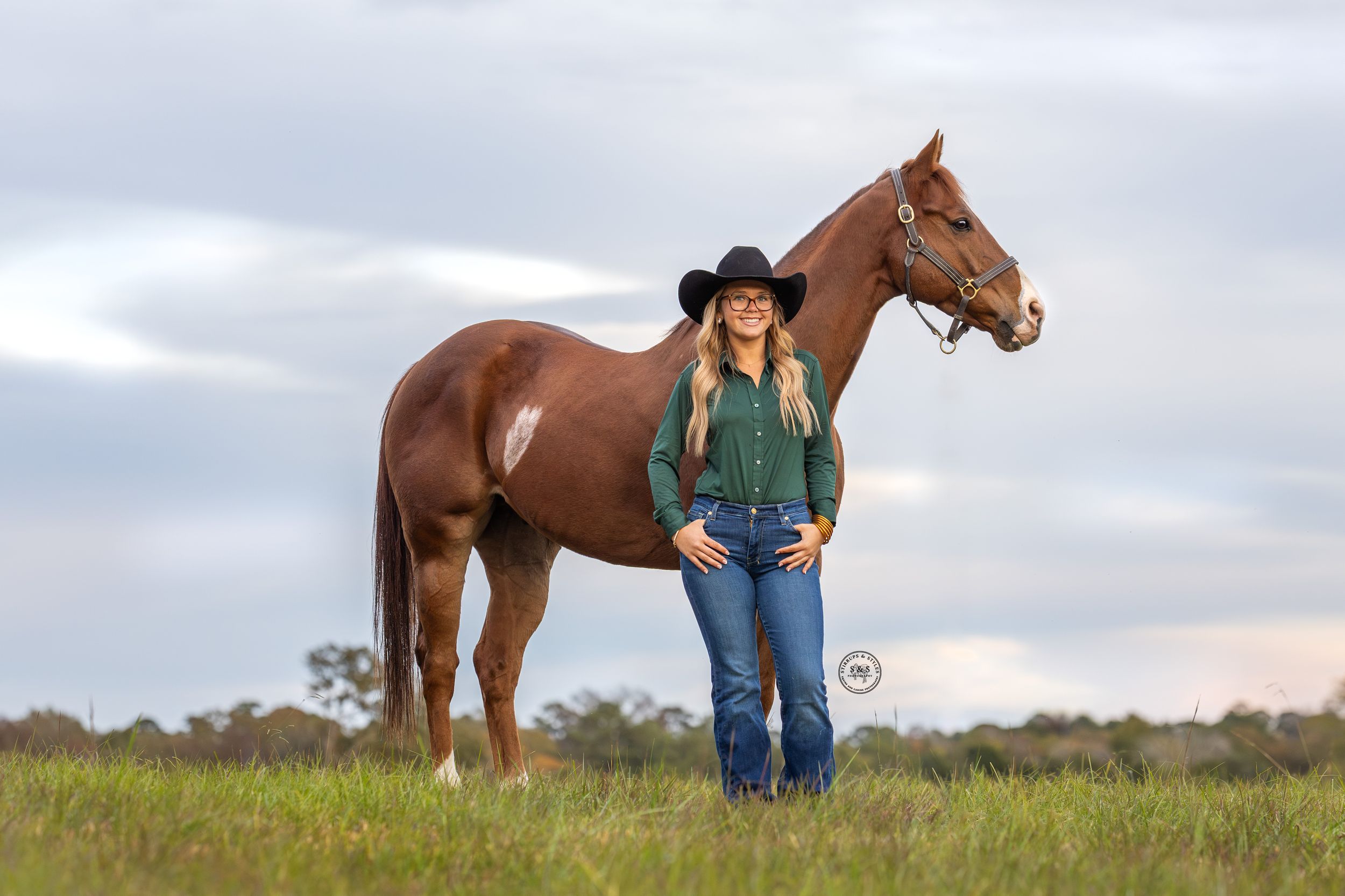 Young woman in black cowboy hat and green shirt stands beside a chestnut horse in a green field.
