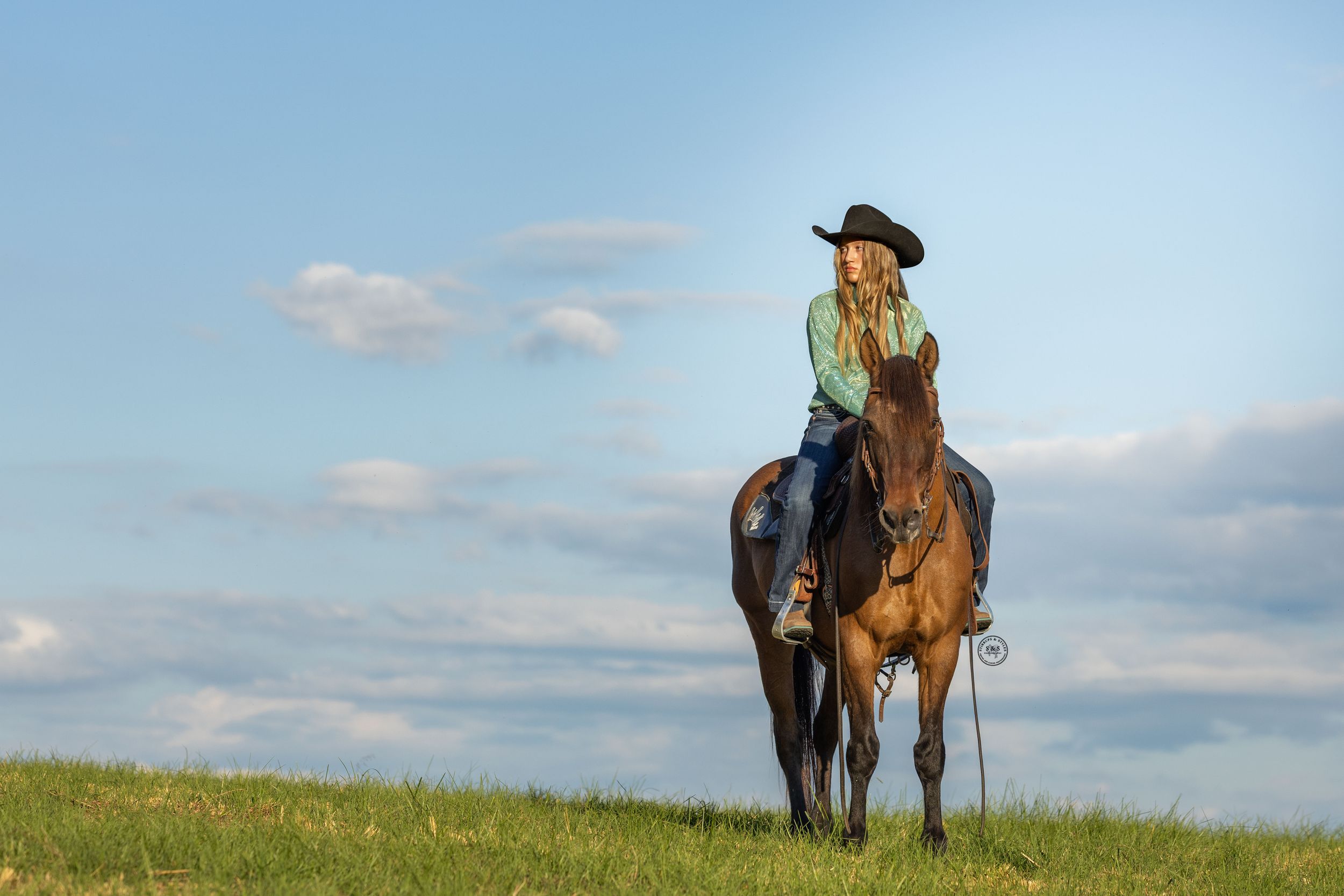 A cowgirl in a black hat rides a bay horse on a grassy hill under a blue sky.