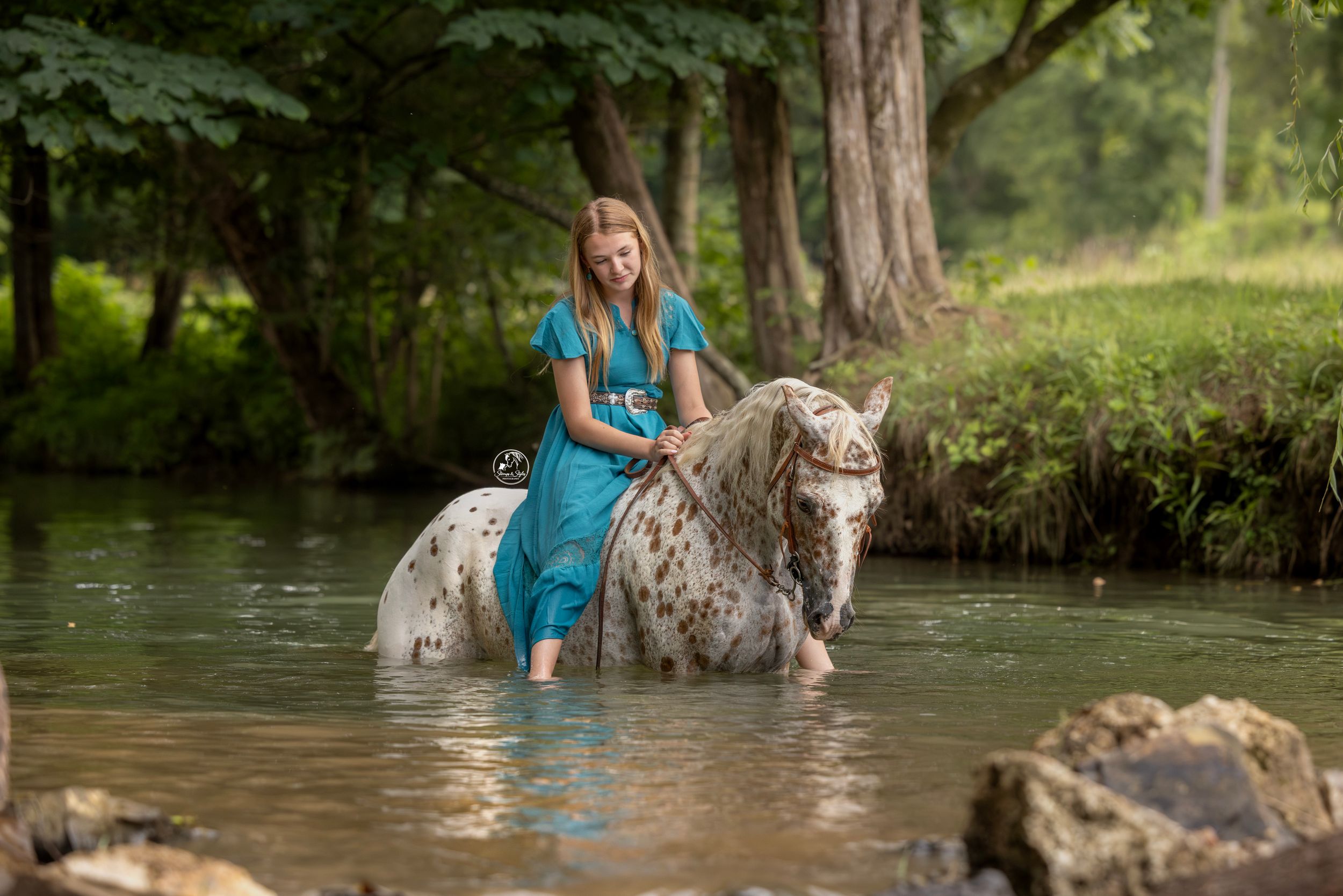 Girl in teal dress riding an Appaloosa horse through a shallow creek surrounded by lush green trees.