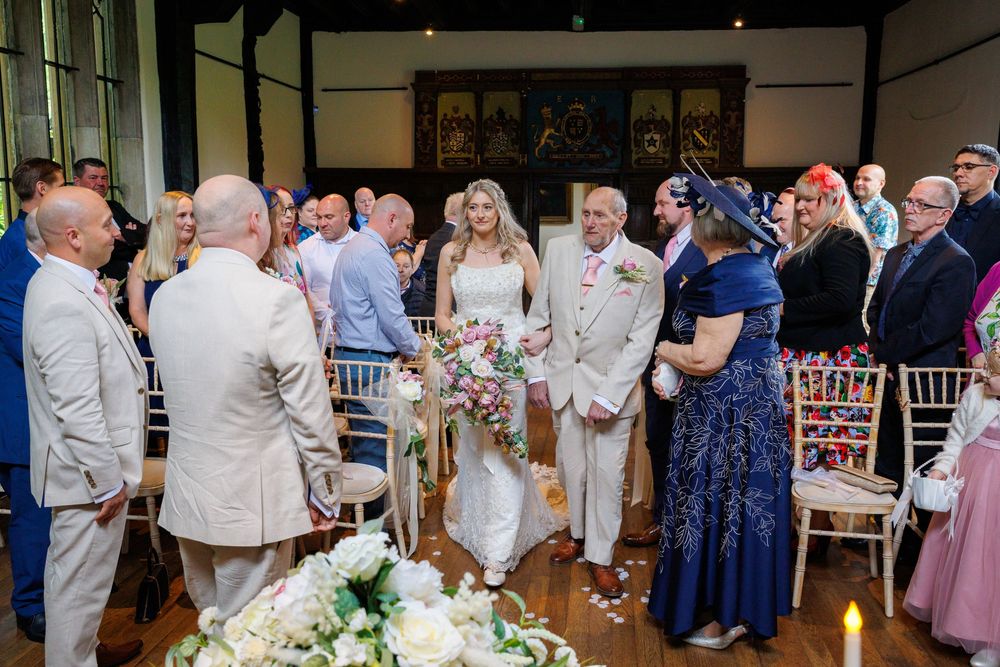 Bride walking down the aisle with escort at elegant indoor wedding ceremony, guests seated on Chiavari chairs.