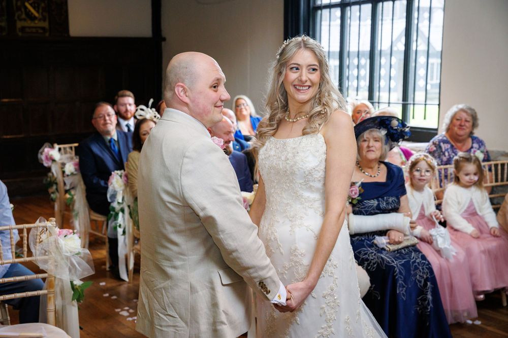 Bride and groom holding hands at wedding ceremony altar, smiling, guests seated in elegant venue background.