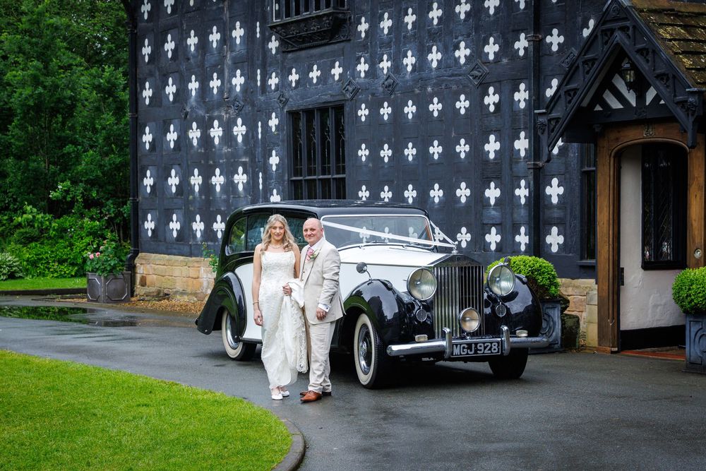 Bride and groom posing beside a vintage Rolls-Royce with white ribbons outside a black and white Tudor wedding venue.