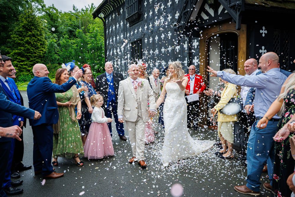 Bride and groom walk through confetti shower outside historic black and white timbered wedding venue surrounded by cheering guests.