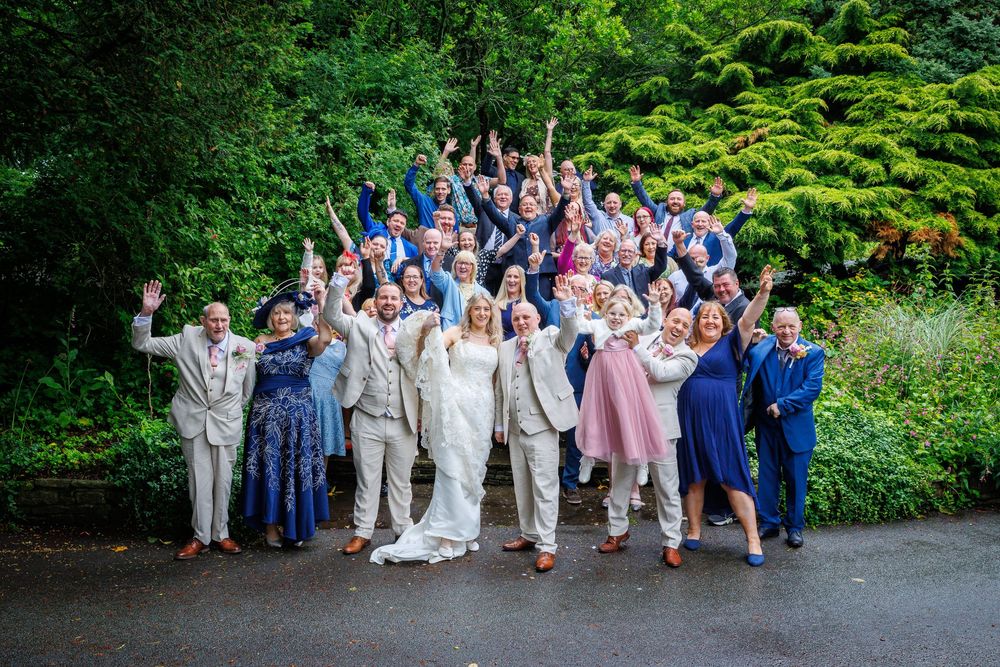 Large wedding party celebrating outdoors on steps surrounded by lush green trees, guests cheering with arms raised.