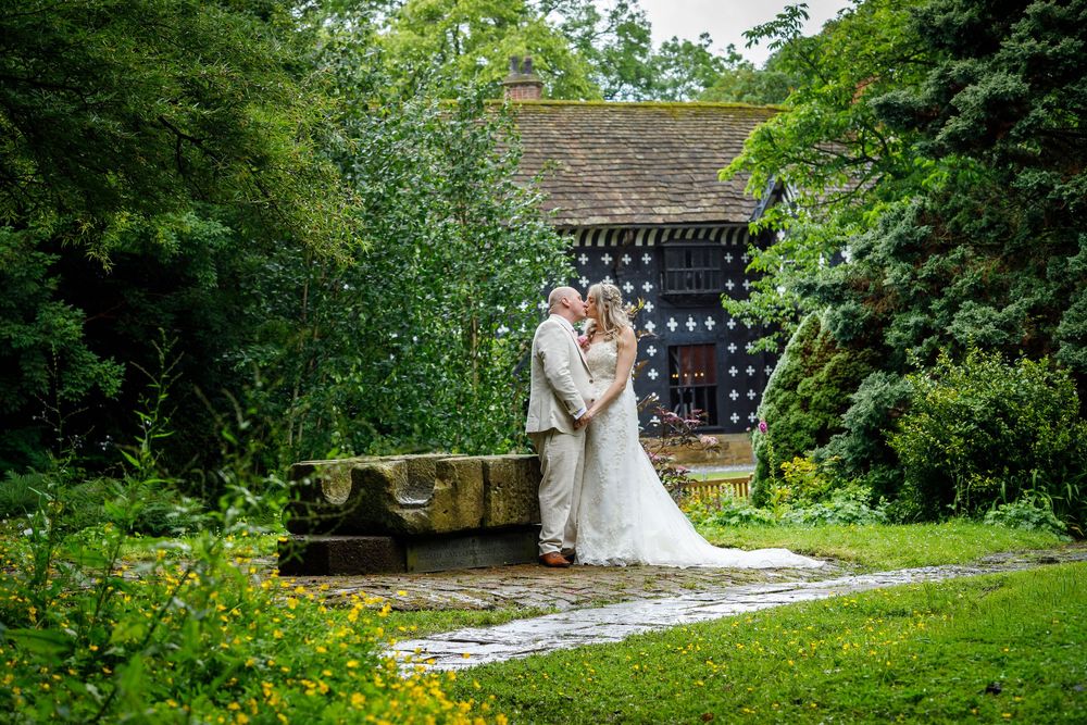 Bride and groom share a romantic kiss in a lush garden setting beside a stone fountain near a charming rustic building.