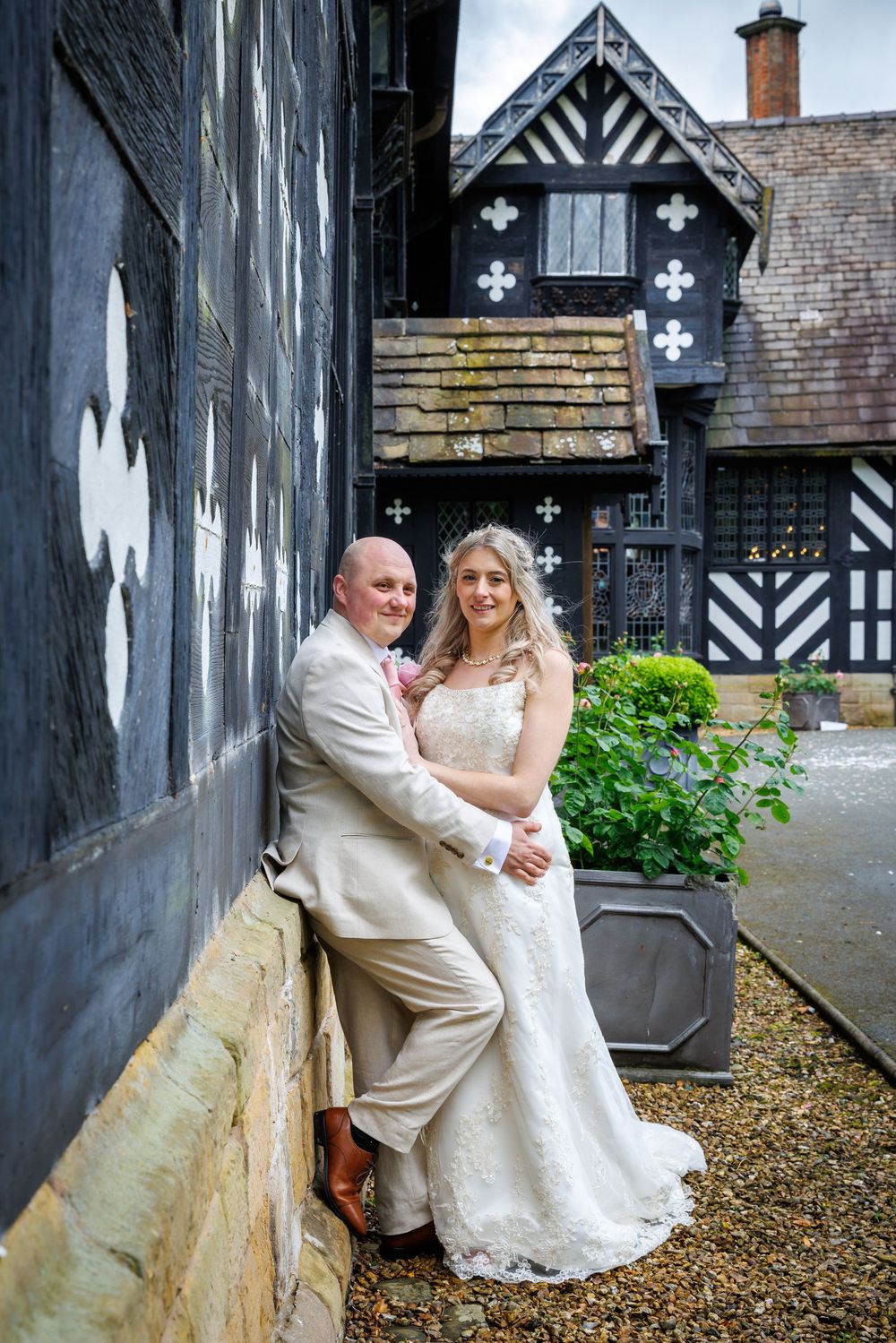 Bride and groom pose together against a black and white Tudor building on their wedding day.