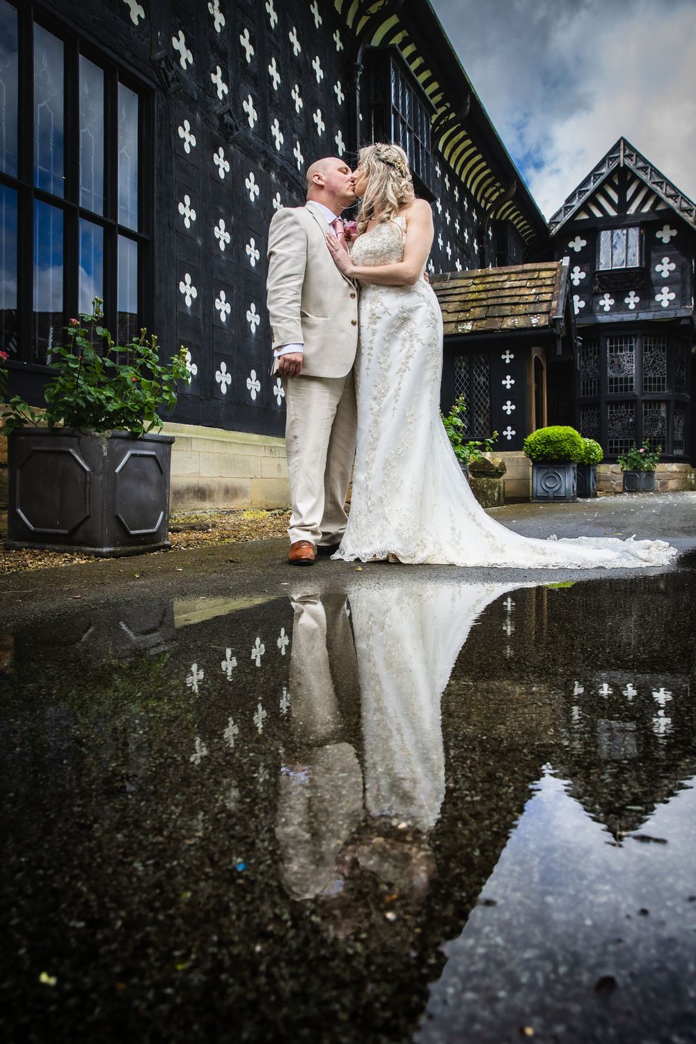 Bride and groom kissing beside a Tudor building, their reflection visible in a puddle below.
