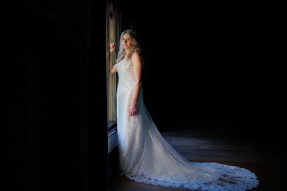 Bride in elegant lace wedding gown with cathedral train stands by dark doorway in dramatic lighting.