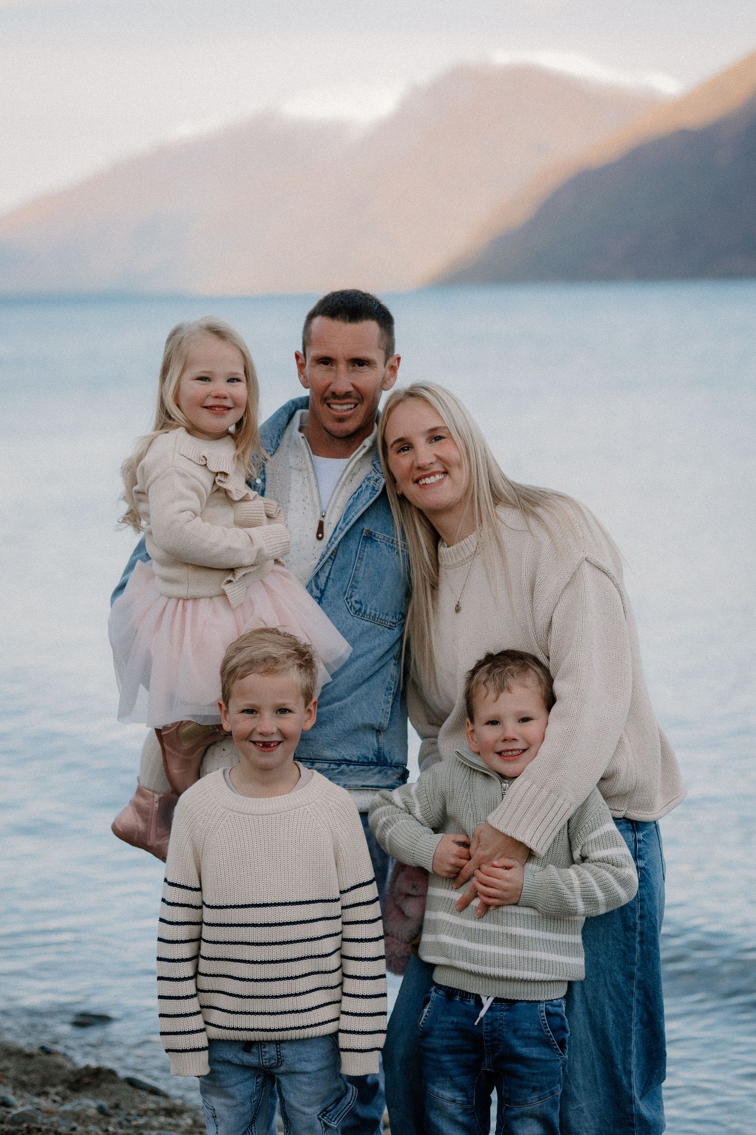 Family of five posing by a mountain lake, parents with three young children in casual knitwear, smiling at camera.