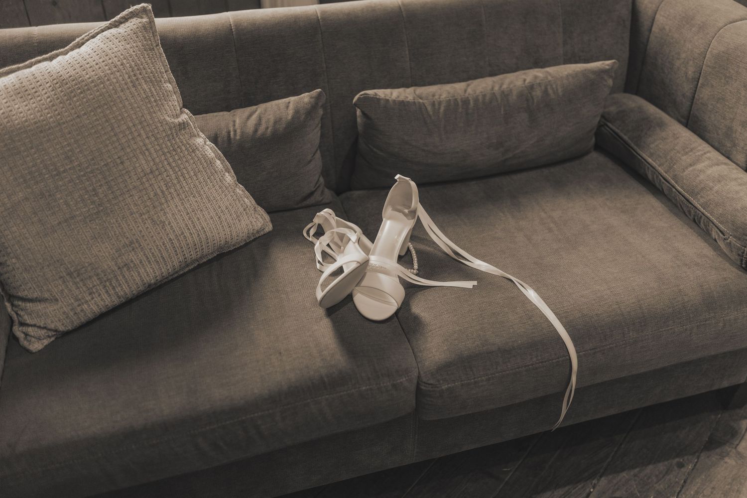 Elegant white heeled sandals placed on a dark gray velvet sofa with a matching cushion.