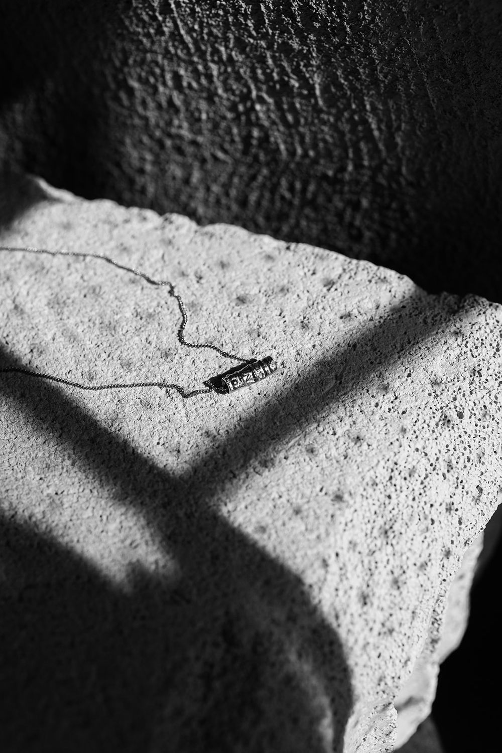 Black and white photo of a delicate bar necklace resting on a textured stone surface with dramatic shadow cross pattern.