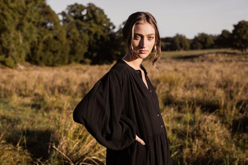 Woman in black dress standing in golden field with trees, outdoor fashion photography in natural light.
