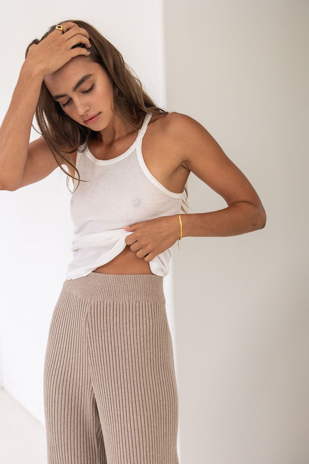 Woman wearing a white ribbed tank top and beige knit wide-leg pants, posing with hand in hair.