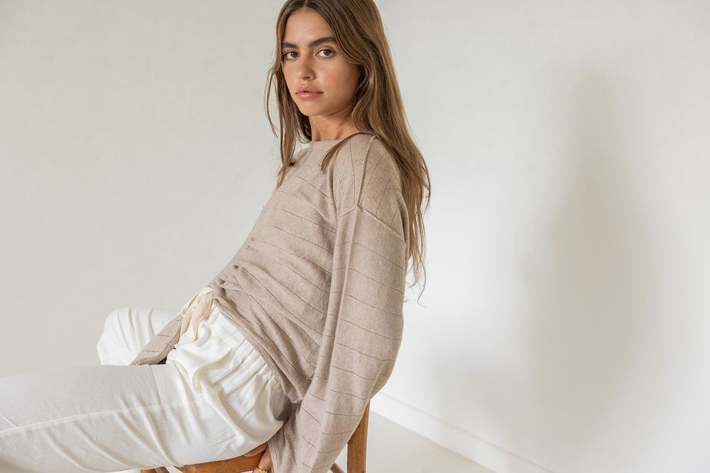 Woman in beige knit sweater and white pants sitting on a wooden chair against a white wall.