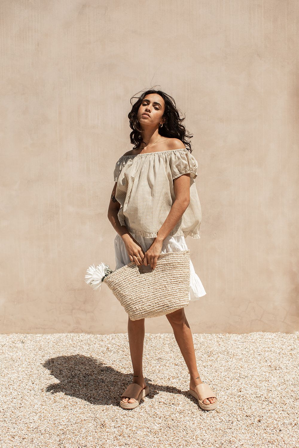 Woman in off-shoulder linen dress holding woven straw tote bag with white flower, wearing sandals outdoors.