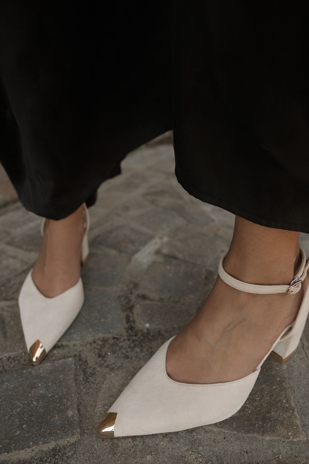 White pointed-toe block heels with gold metal cap toe and ankle strap on cobblestone, paired with black wide-leg trousers.