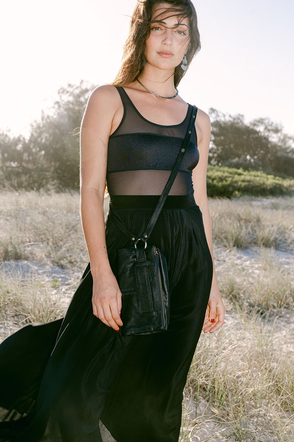 Woman in black mesh bodysuit and maxi skirt holding crossbody bag outdoors in natural setting.